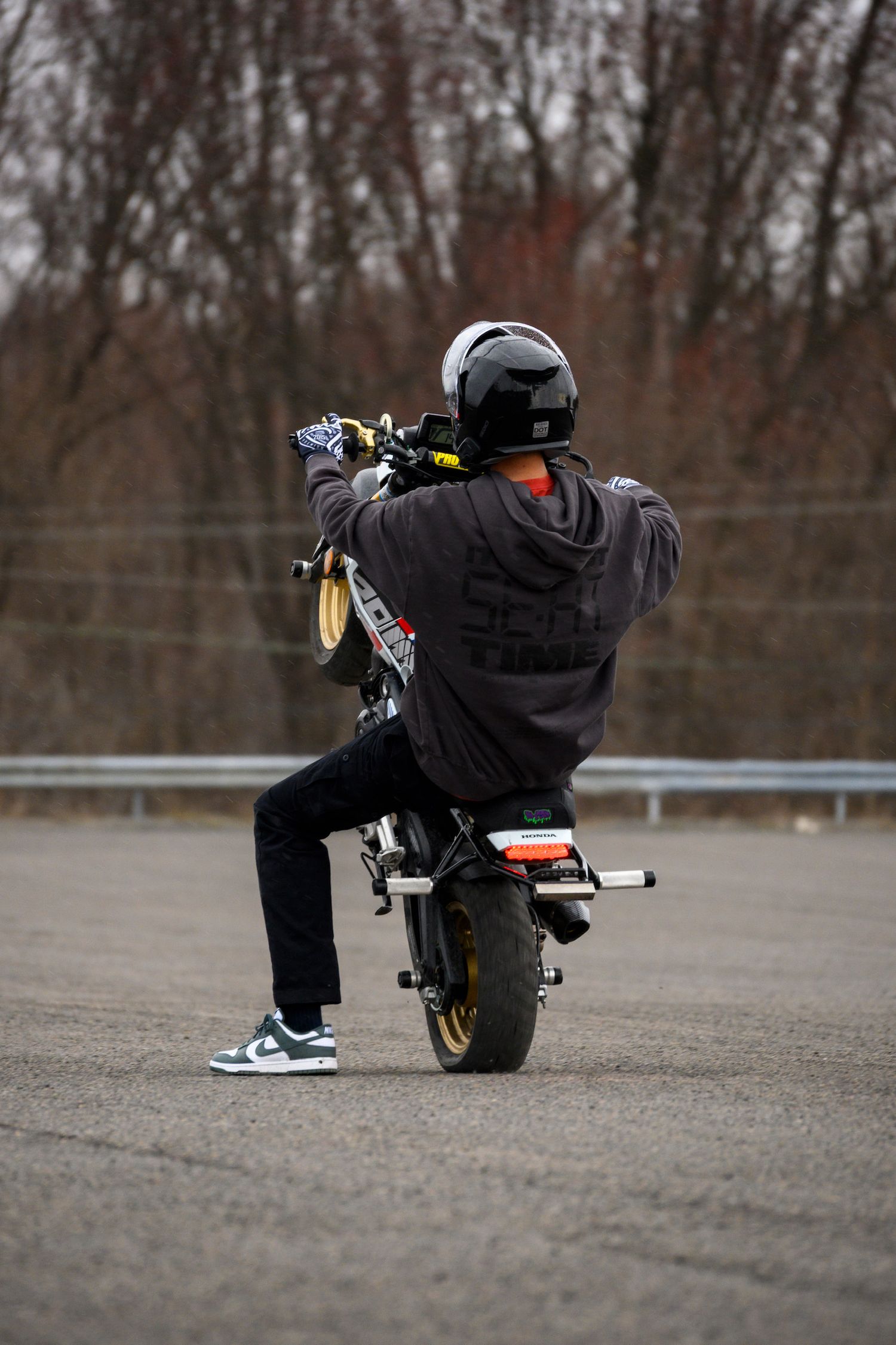Motorcyclist performing a stunt on a small bike in an empty parking lot, wearing a black helmet and hoodie.