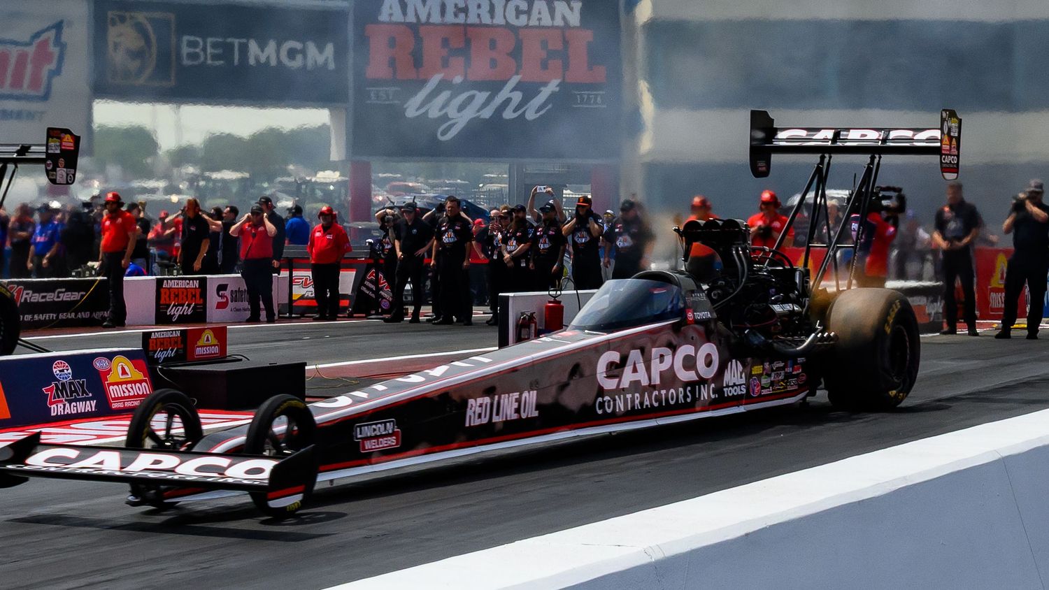 A red CAPCO dragster racing down the track with spectators in the background.