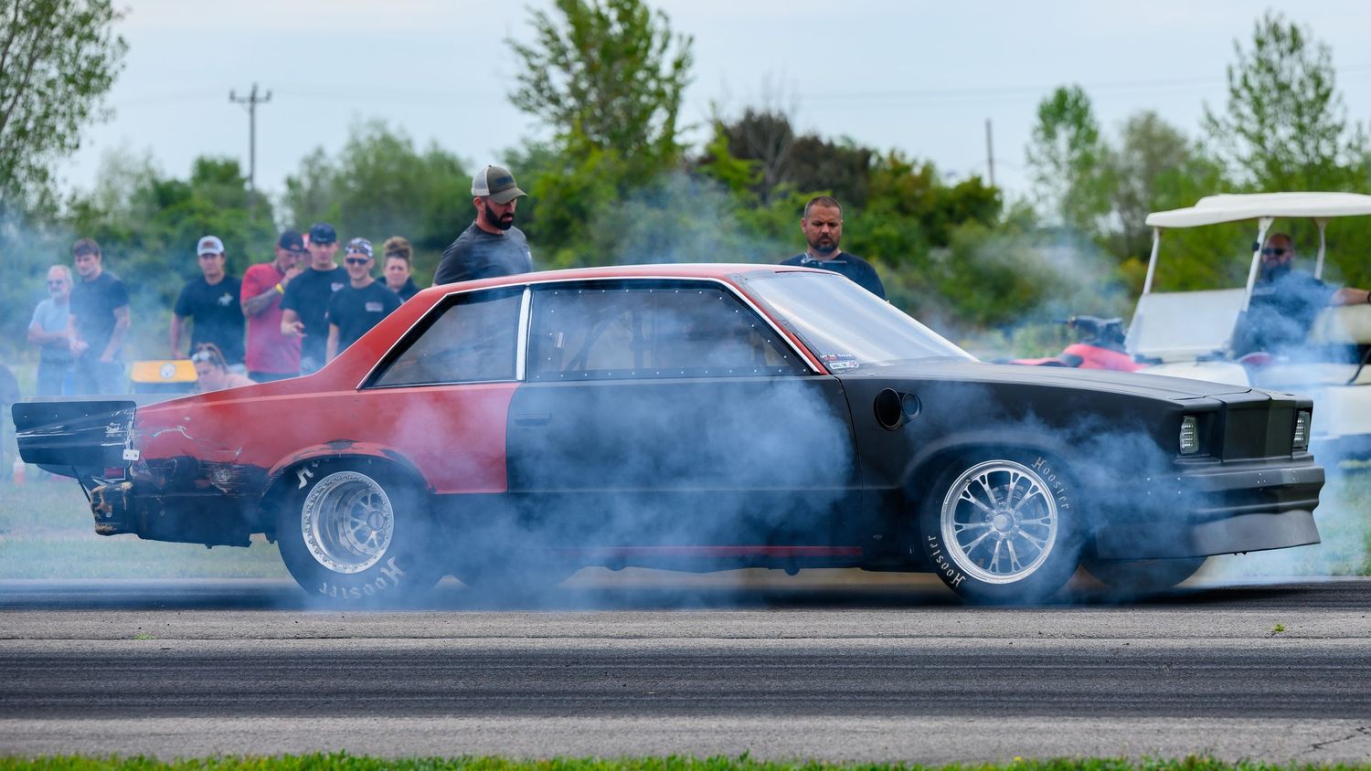 Multiple dark sports cars creating tire smoke during a drift event on a sunny day.