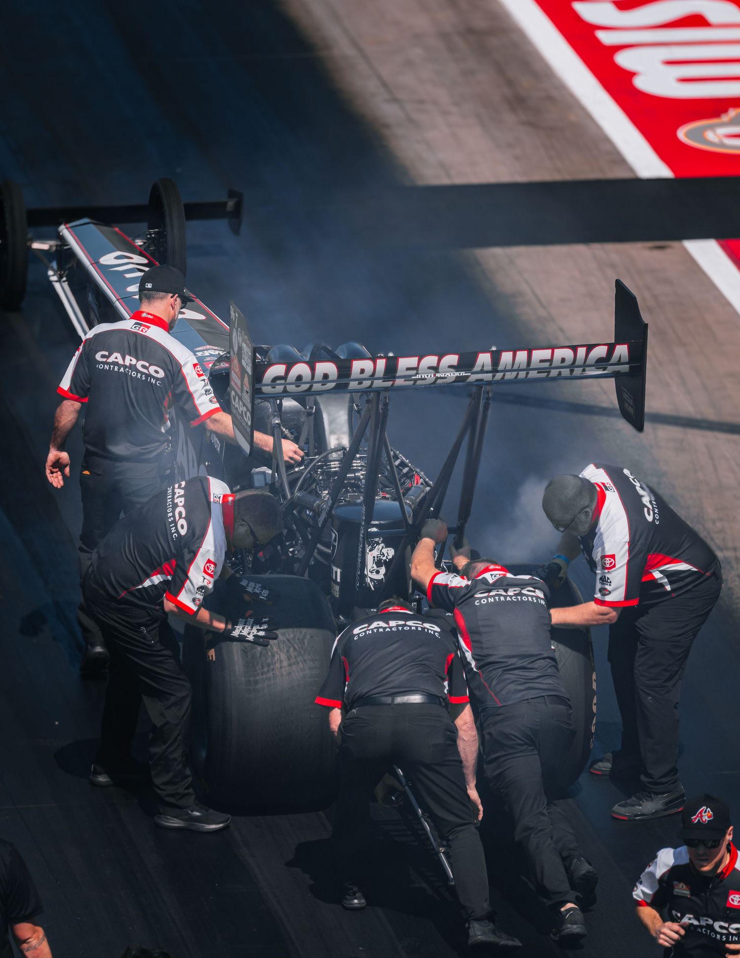 Racing pit crew working on a dragster with 'God Bless America' text visible on the wing.