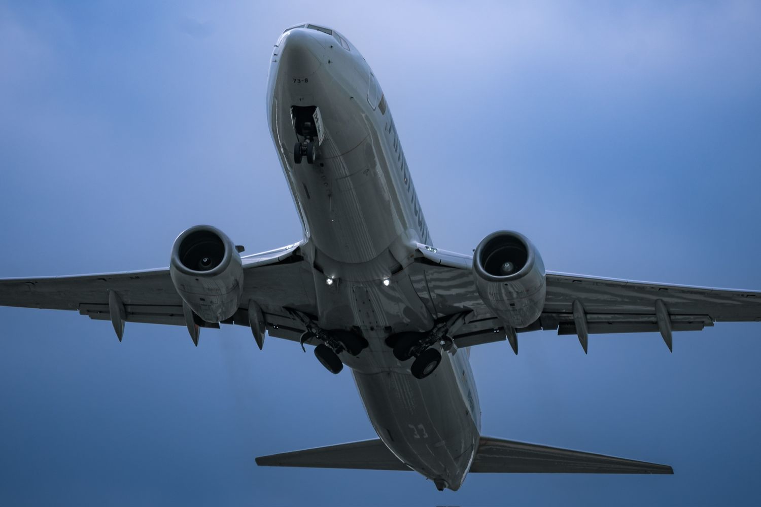 Commercial airplane ascending against a dramatic blue sky, viewed from below showing twin engines and landing gear.