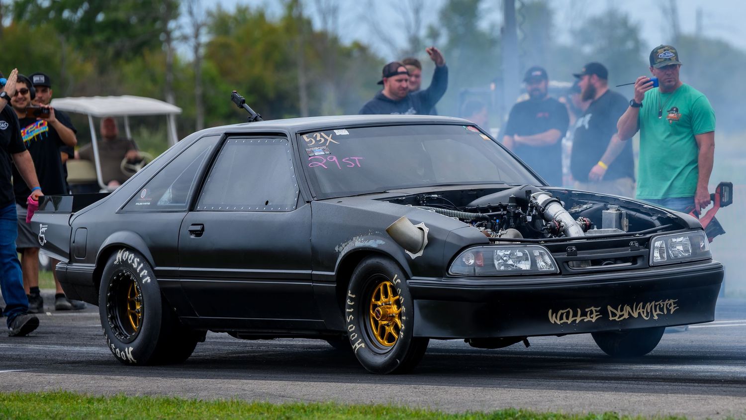 Classic cars line up at a drag racing event with smoke and burnouts on the track.