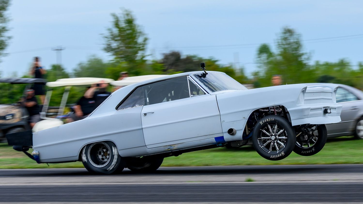 Modified vintage cars racing down a drag strip with visible tire smoke.