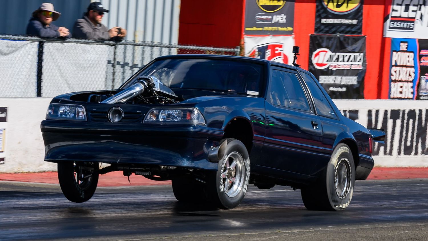 Dark colored muscle cars competing in a drag race on a professional track.