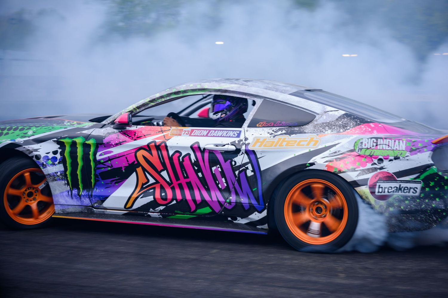 A colorful Monster Energy drift car with orange wheels spins on a track, creating dramatic smoke clouds during a racing event.