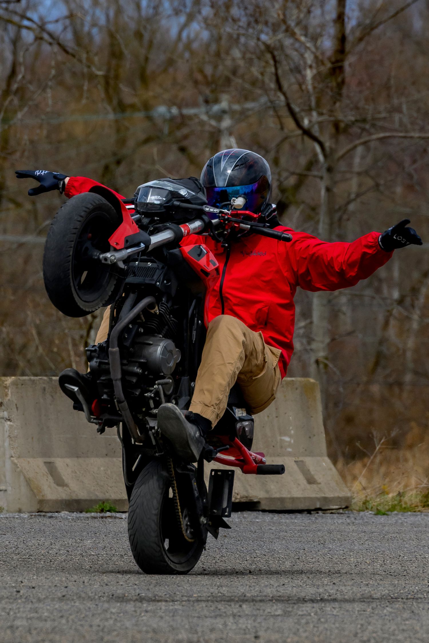 Motorcyclist performing a wheelie stunt in a red jacket and helmet on an outdoor road.
