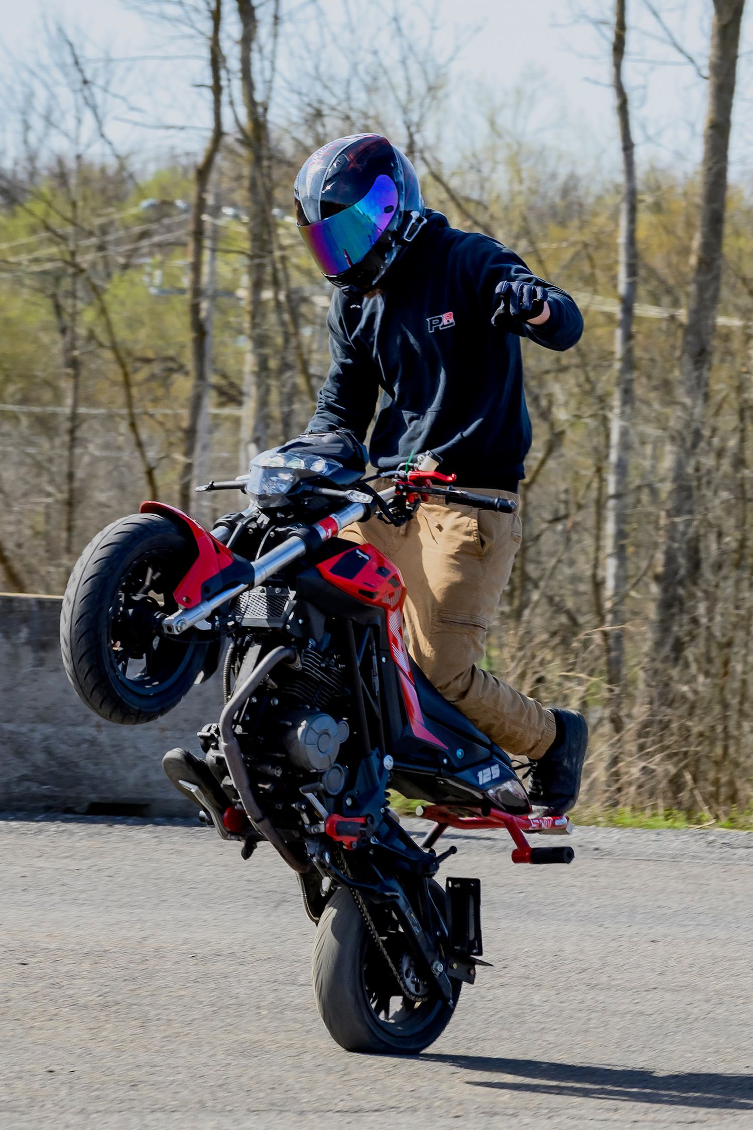 Motorcyclist performing a wheelie stunt on a red and black bike while wearing a helmet and casual clothing outdoors.