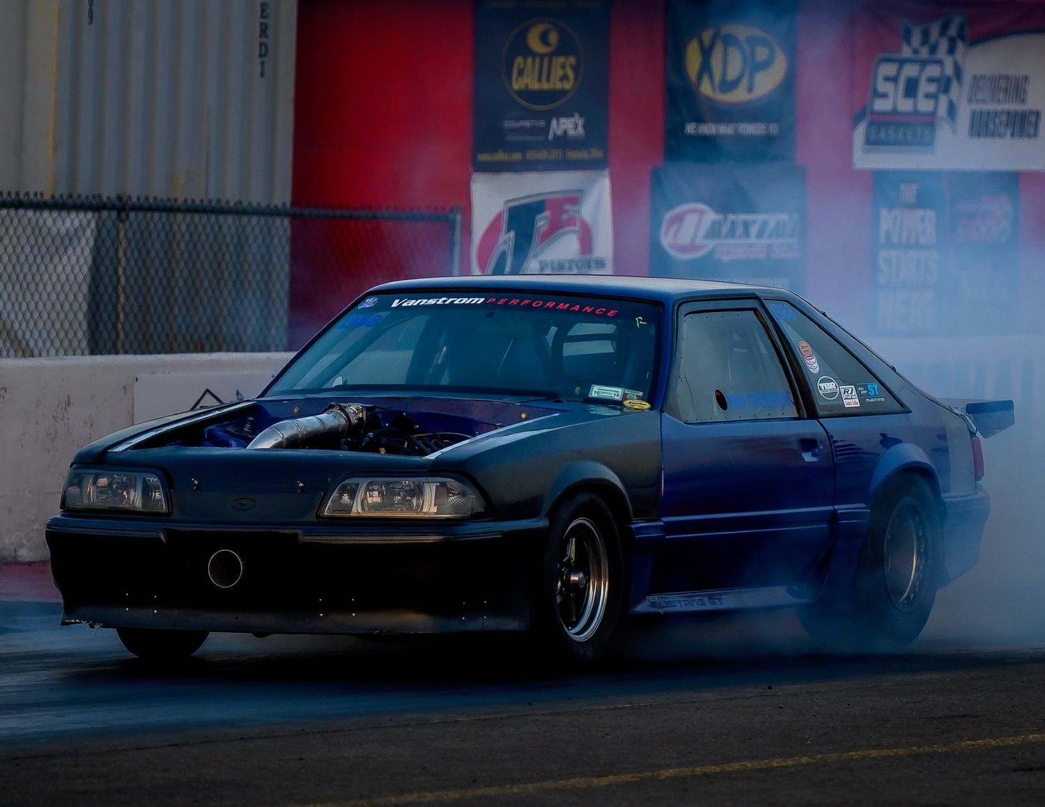 A dark blue classic car drifts around a corner with smoke billowing from the tires during a drift competition.