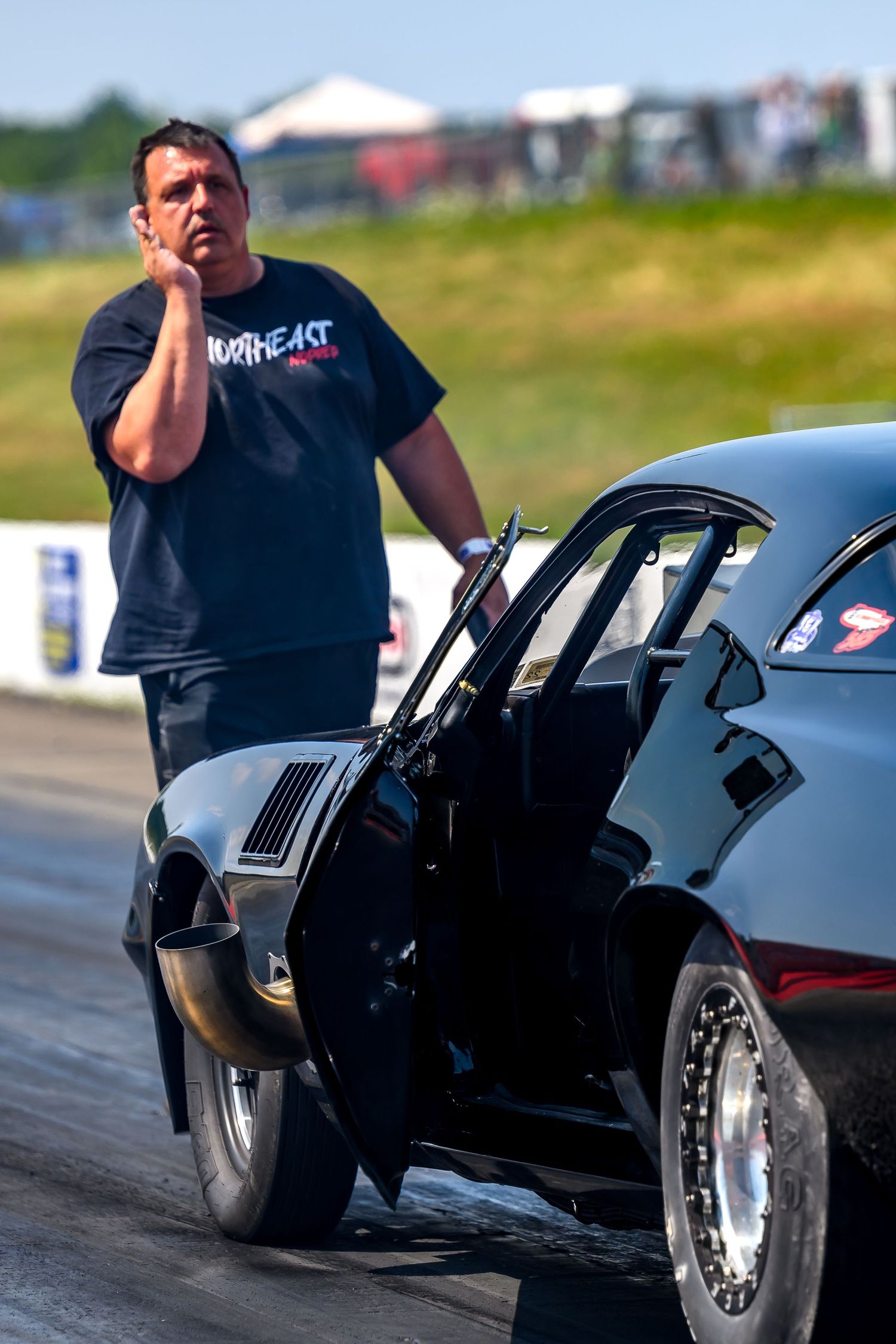 A person in a black t-shirt crouches next to a dark custom drag racing car on a track.