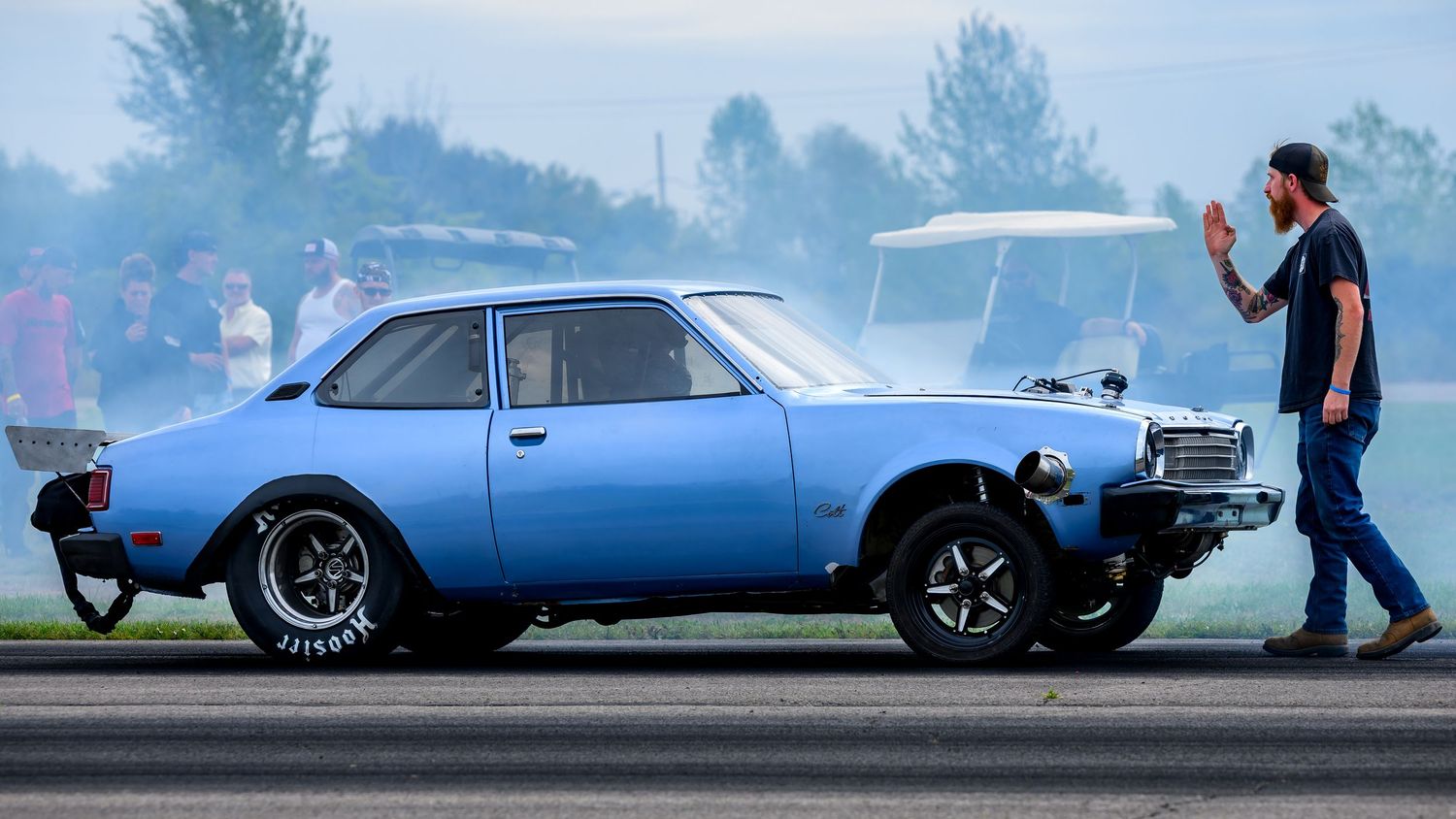 A blue classic car with lifted front wheels racing on a drag strip.