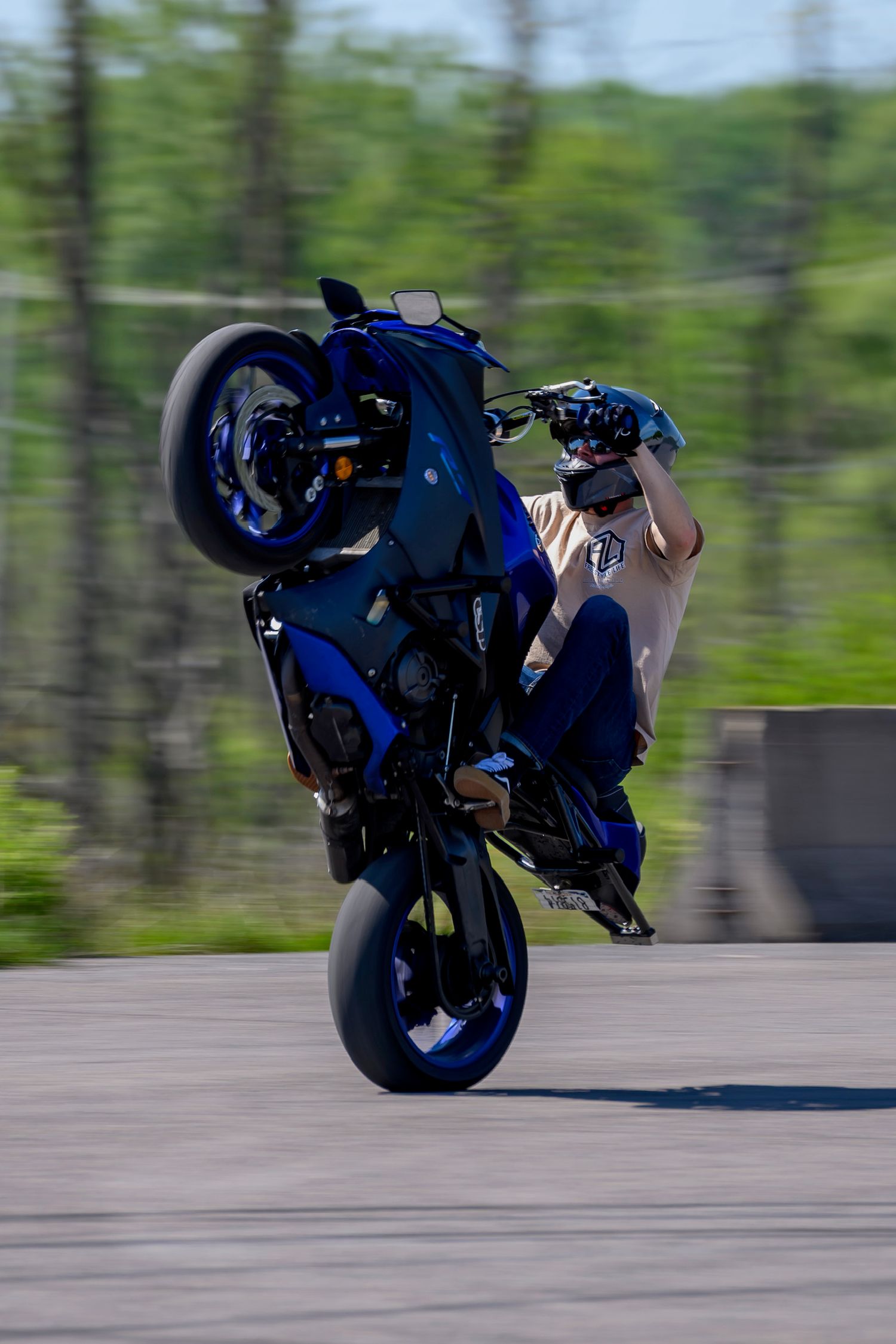Motorcyclist performing a wheelie stunt on a blue sport bike on a paved track with green trees in the background.