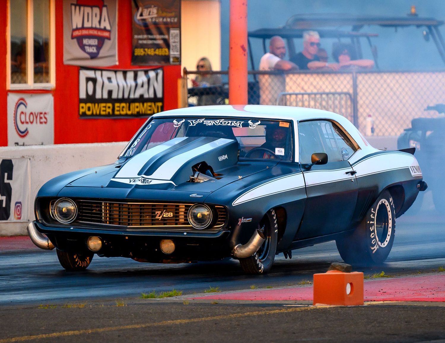 A classic drag racing car launches from the starting line with visible sponsor signage.