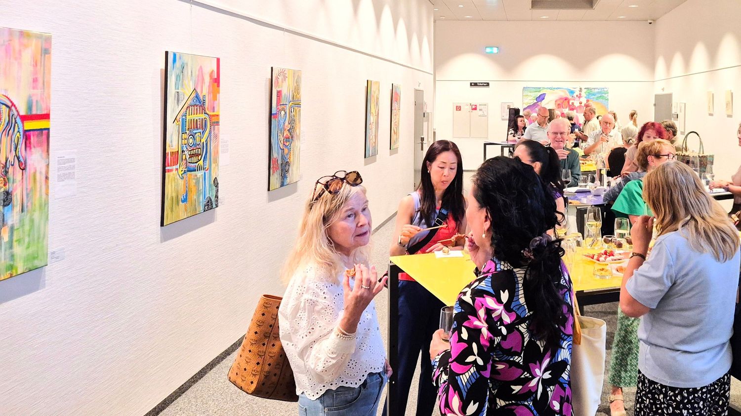 People mingling at a colorful art gallery exhibition with vibrant paintings displayed on white walls.