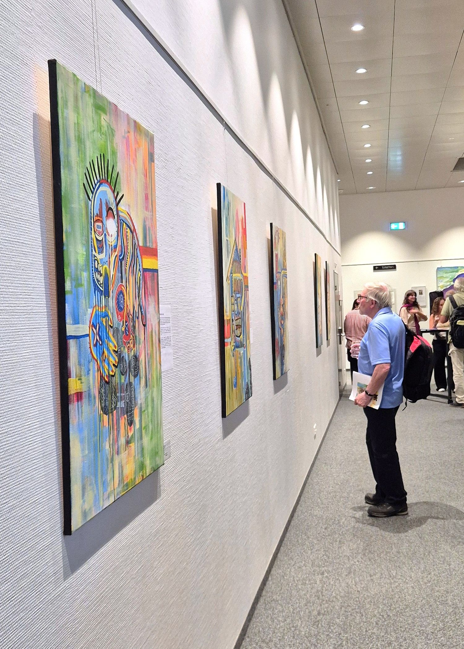Visitors viewing colorful contemporary paintings displayed on white gallery walls in a modern art exhibition hallway.
