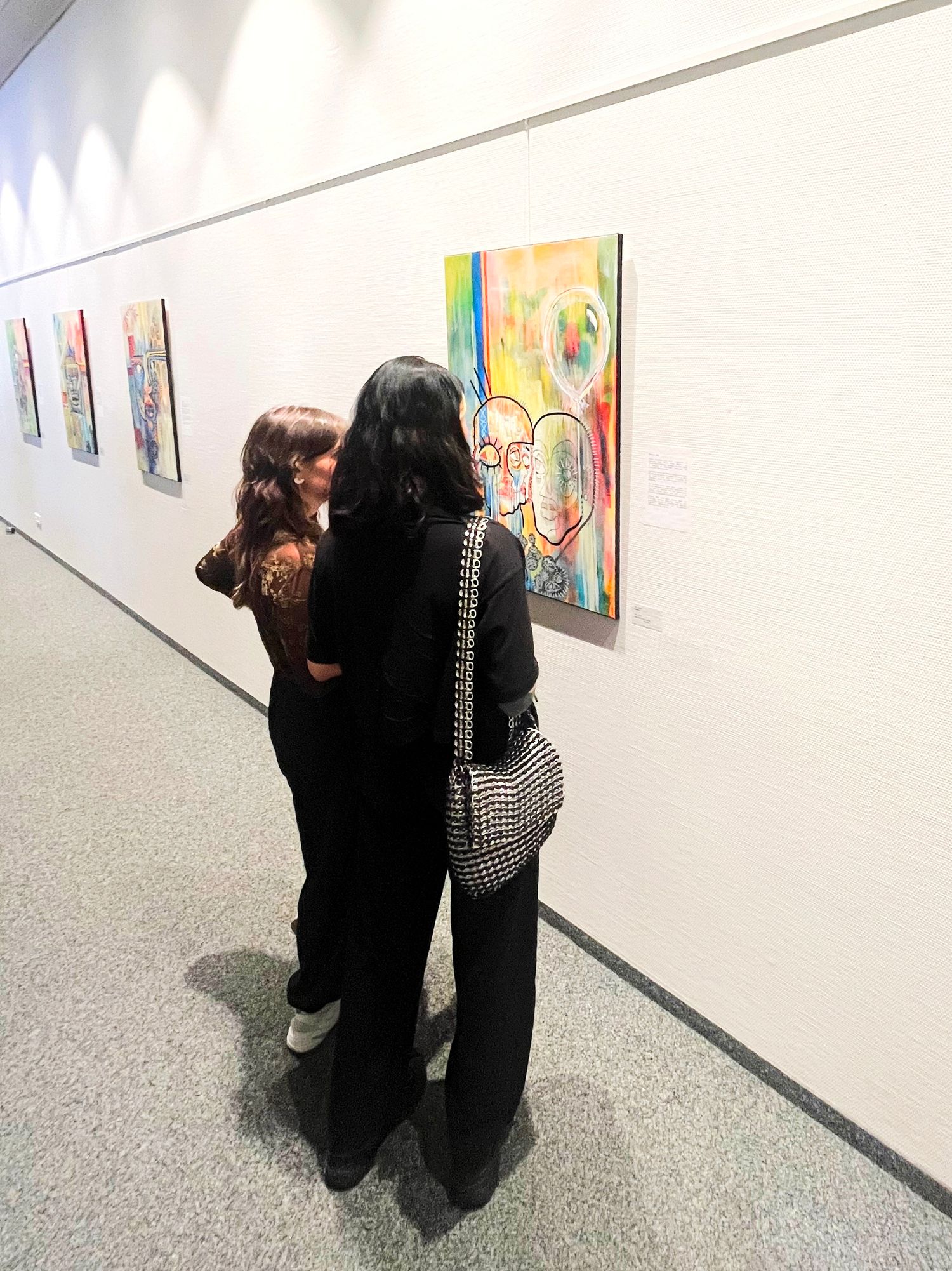 Two women viewing colorful abstract paintings on a white gallery wall during an art exhibition.