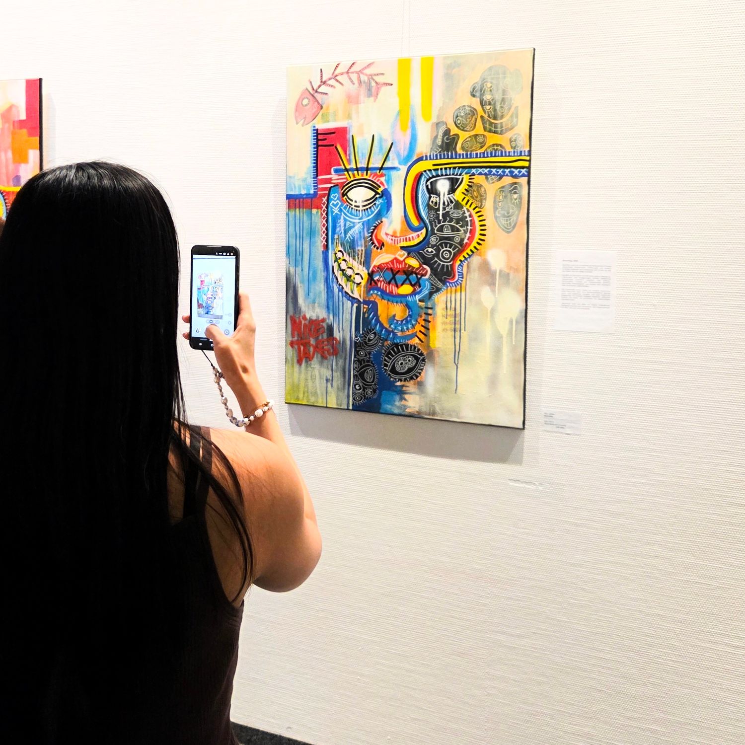 Woman photographing colorful abstract art painting on white gallery wall with smartphone.