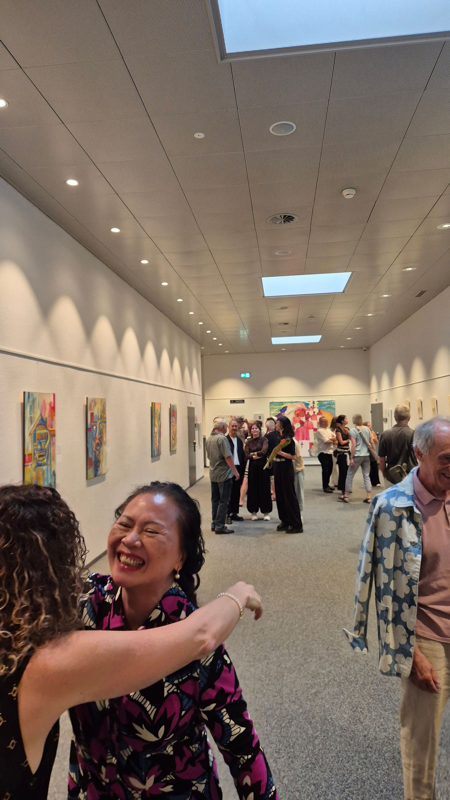 Guests laughing and socializing at an art gallery opening with colorful paintings displayed on white walls.