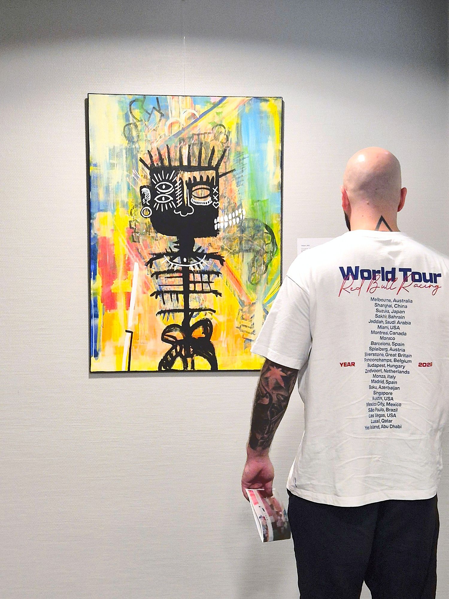 Tattooed man in white World Tour t-shirt viewing colorful Zailou artwork on gallery wall.