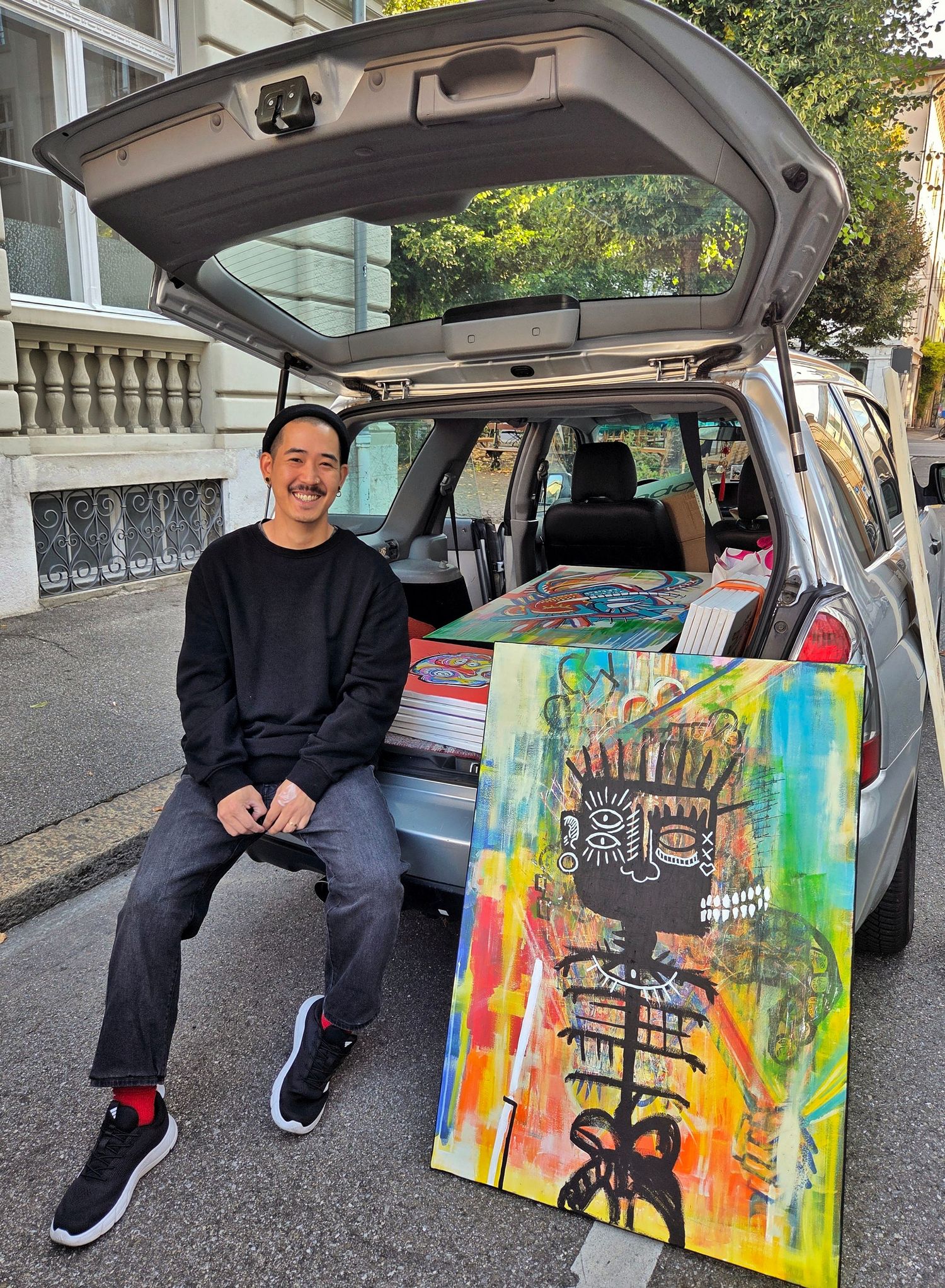 Zailou smiling beside colorful abstract paintings in open car trunk on urban street.