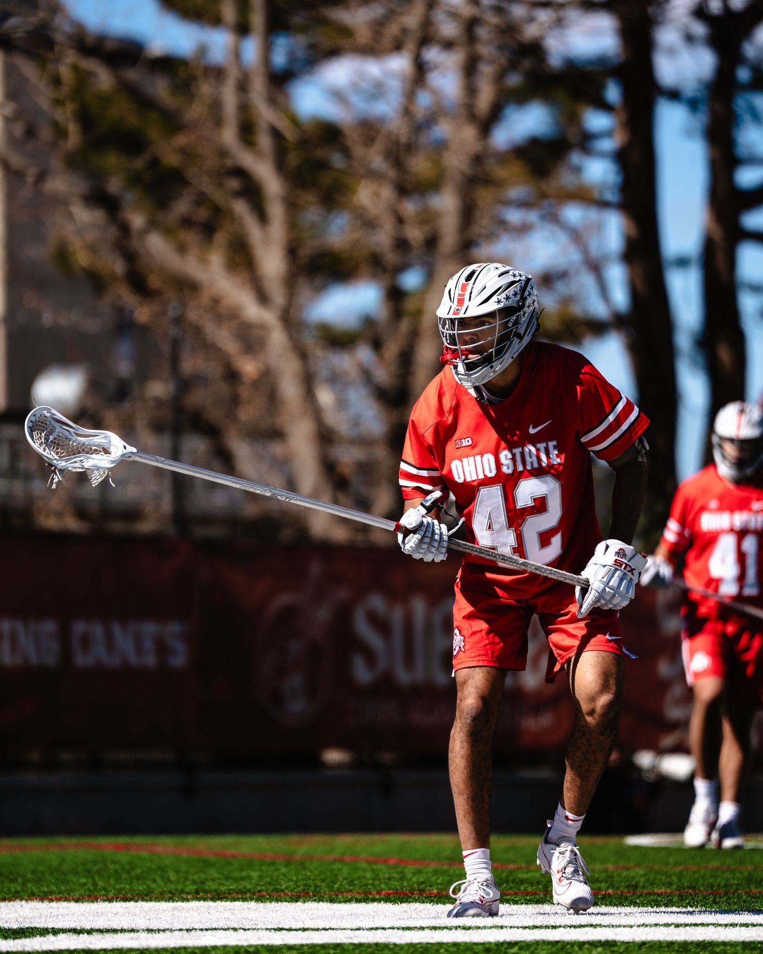 Ohio State lacrosse player in red jersey crouching with stick during game action.