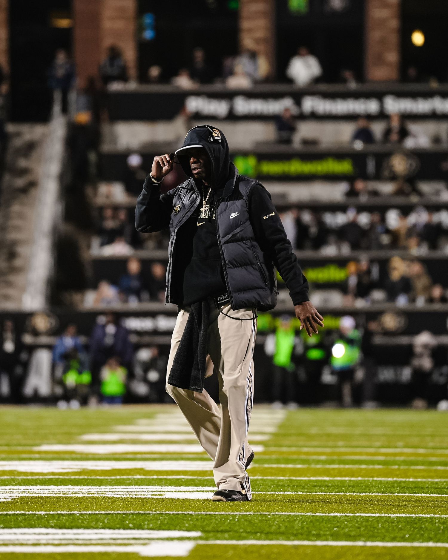 Football coach in black puffer jacket walks across field during nighttime game.