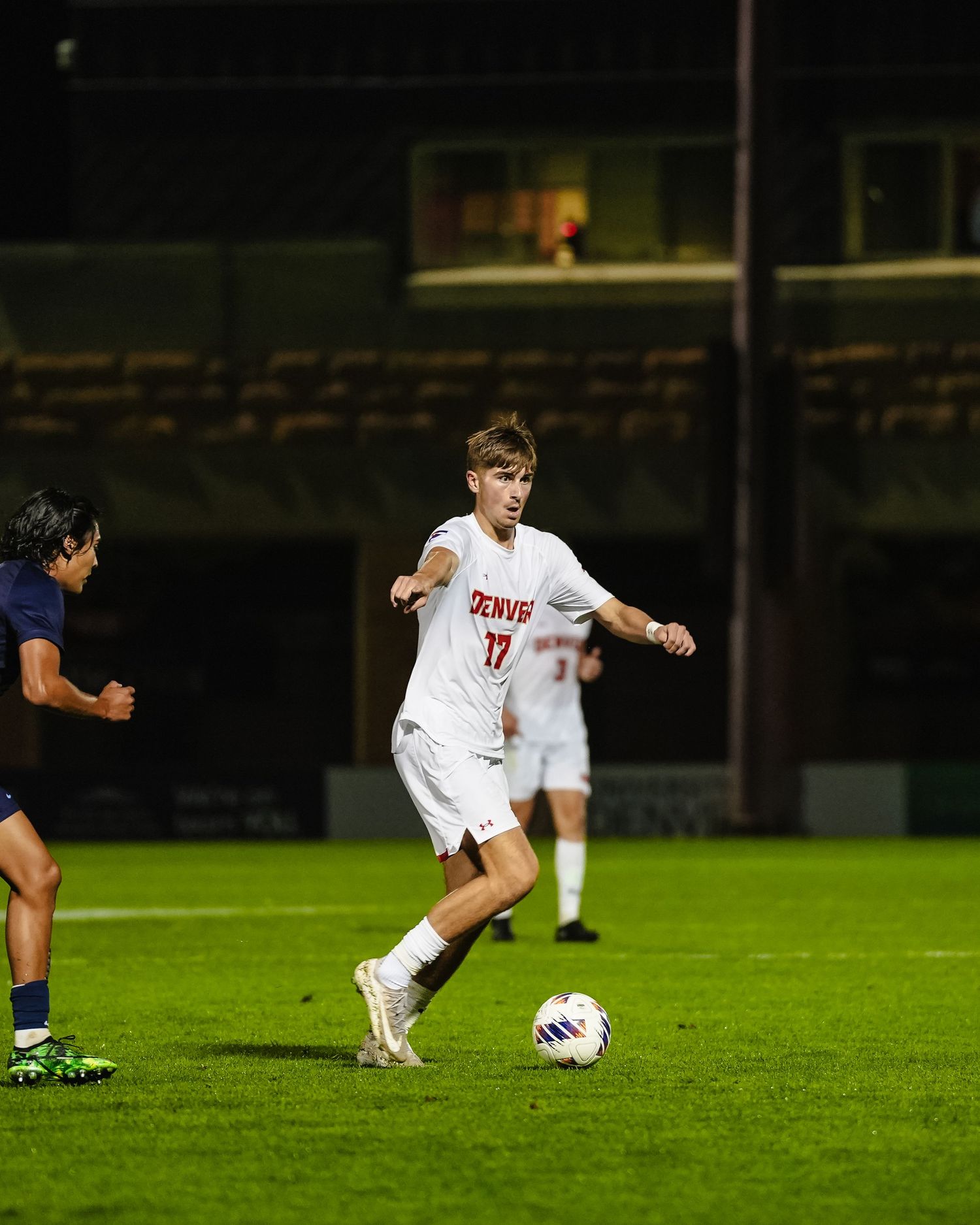 Soccer player in white uniform dribbling ball across illuminated field at night.