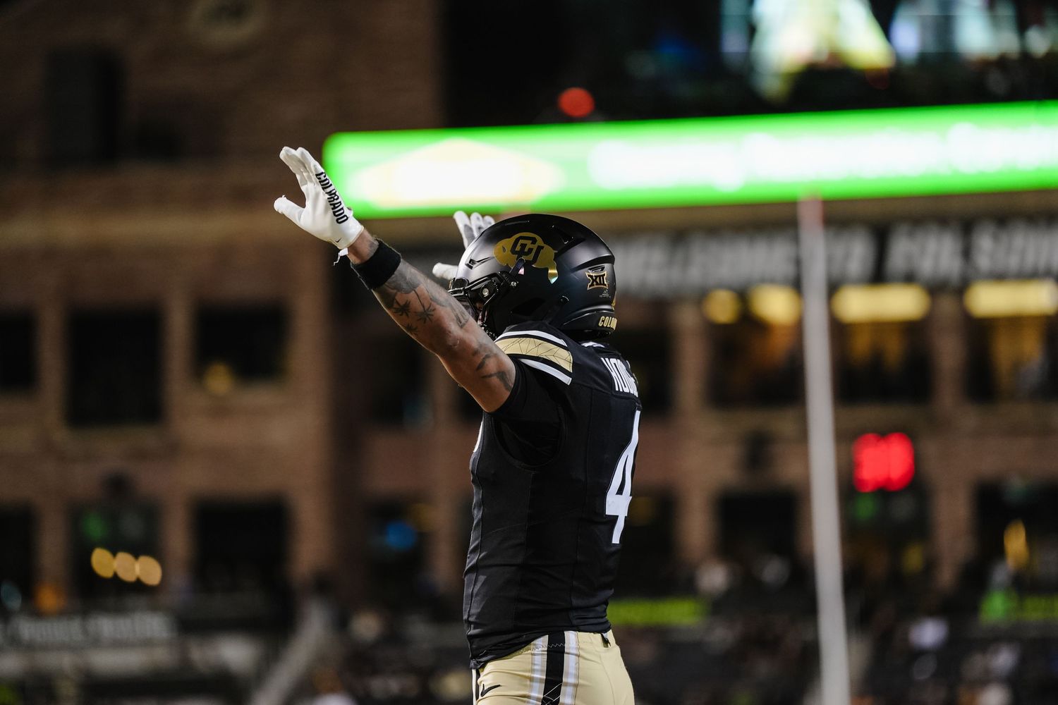 Football player in black uniform raises arms on field during night game.