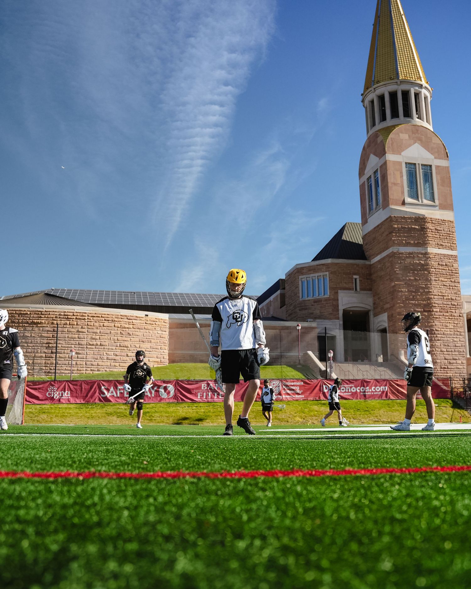 Lacrosse players practicing on field with brick building and blue sky in background.