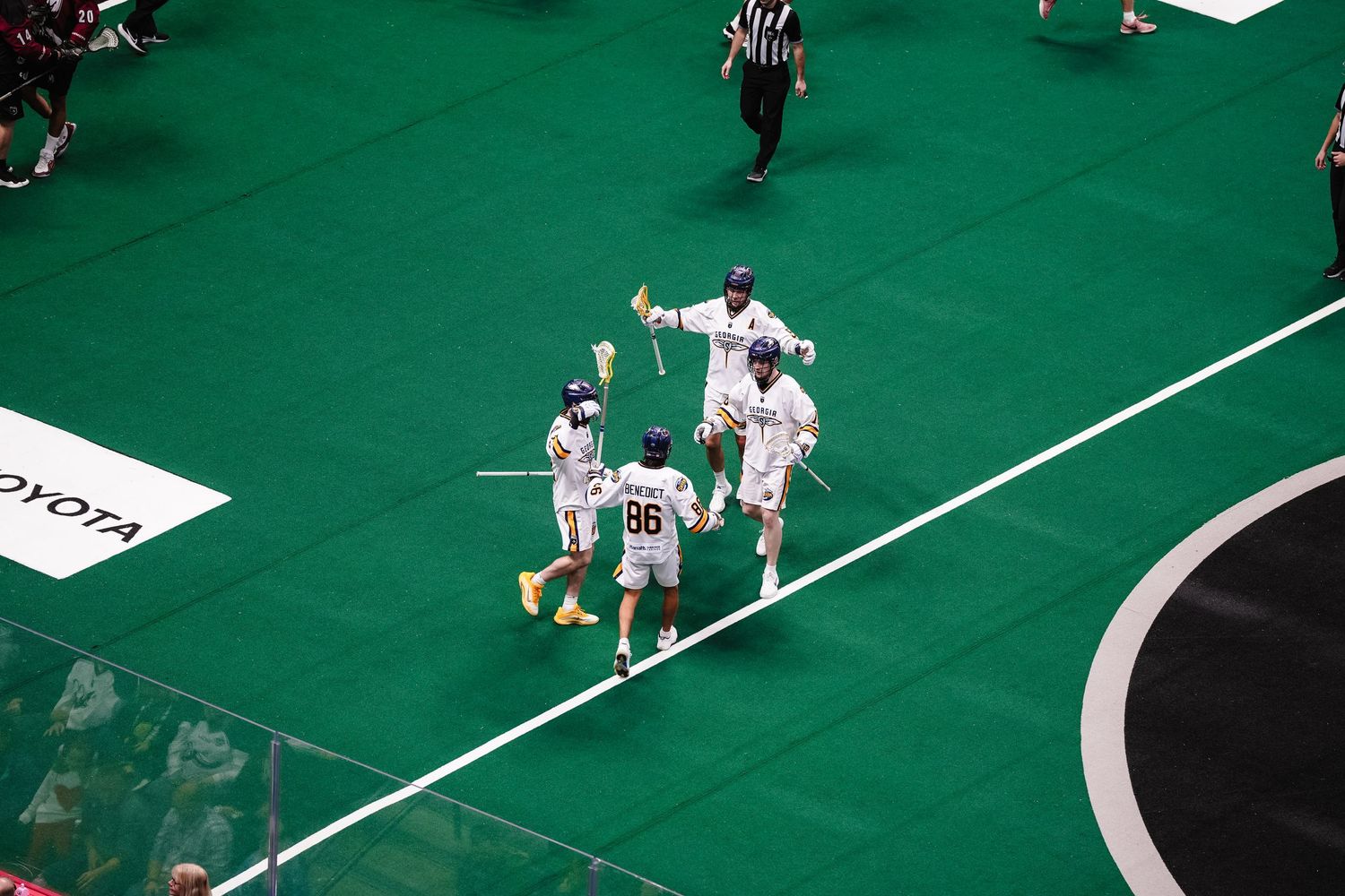 Players competing on bright green turf field during indoor lacrosse match.