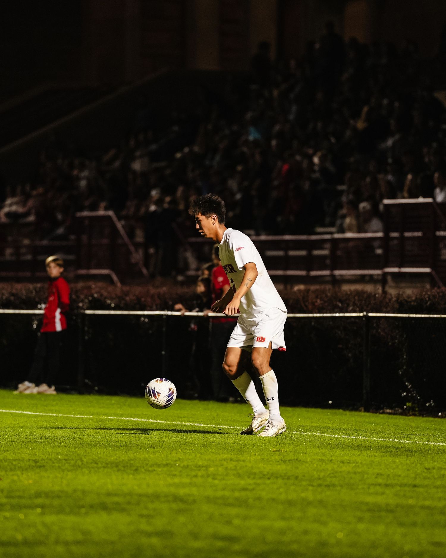 Player in white uniform controls ball on illuminated lacrosse field at night.