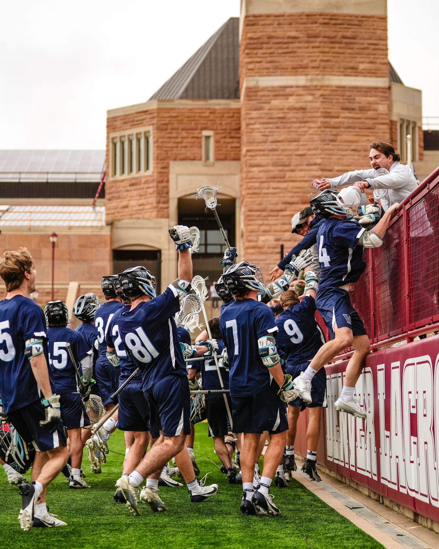 Lacrosse team in navy uniforms celebrating with fans at stadium fence.