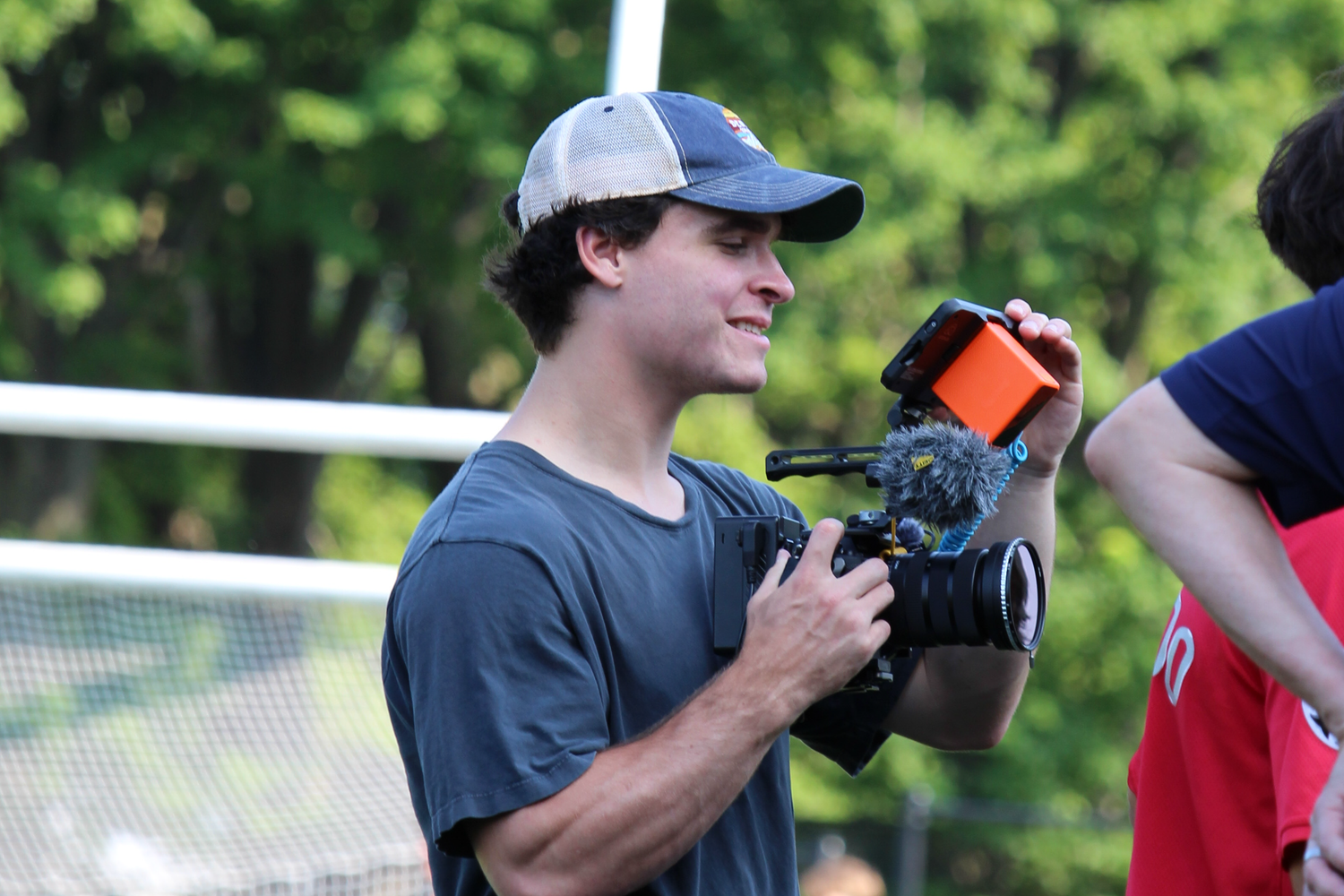 Camera operator filming sports event while wearing baseball cap.