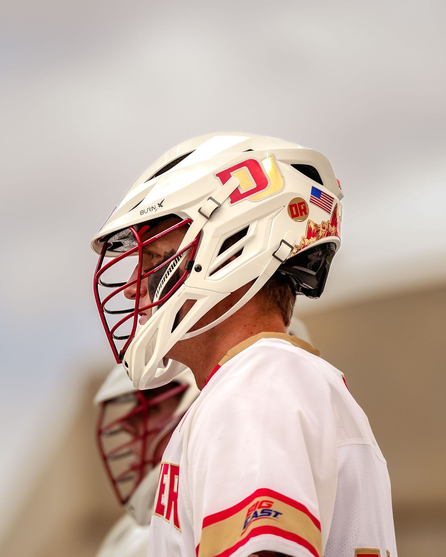 Lacrosse player wearing white jersey and helmet with red accents during a game.