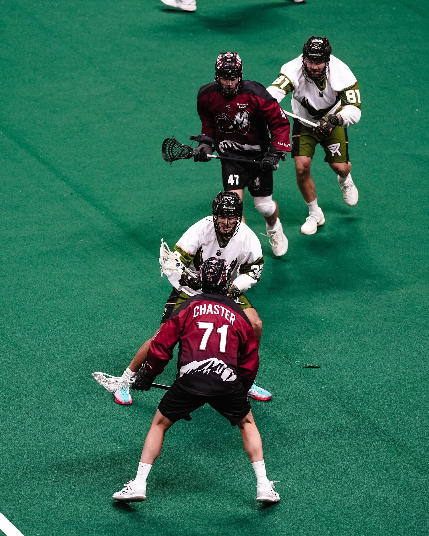 Indoor lacrosse players compete on a bright green turf field during a professional match.