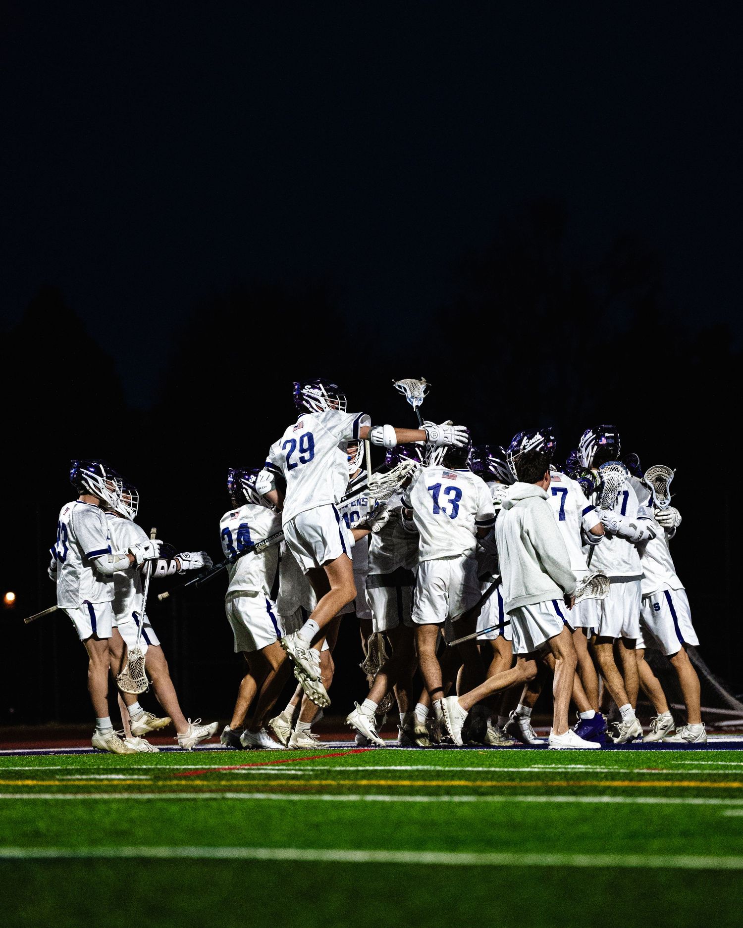 Team of lacrosse players in white uniforms celebrating together on a field at night.
