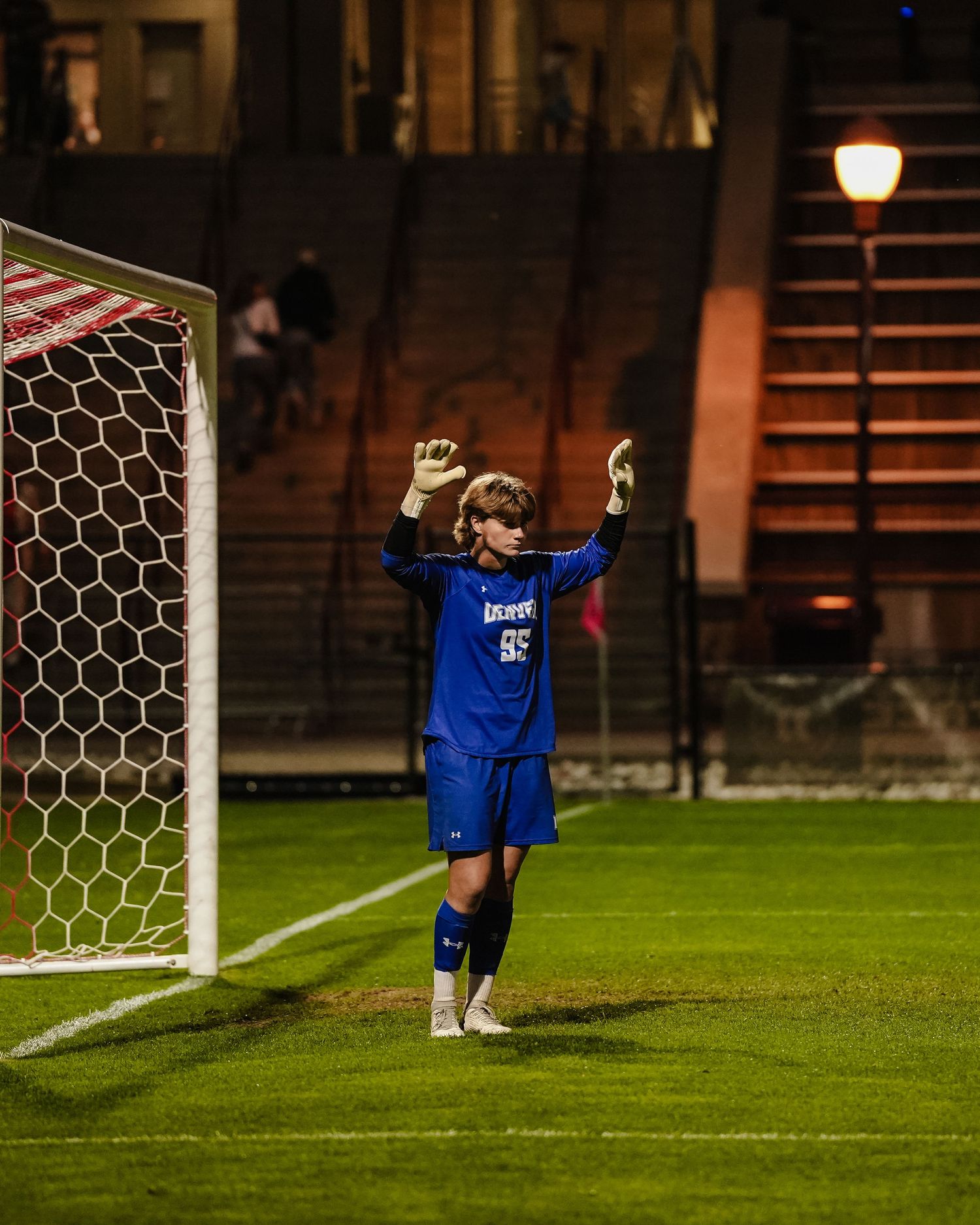 Soccer goalkeeper in blue uniform stretching arms wide in front of goal net at night.
