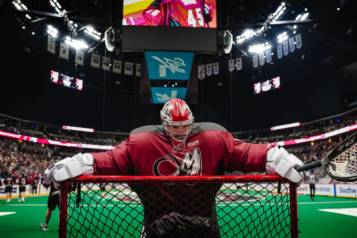 Lacrosse goalie in red uniform stretches across goal during indoor game.