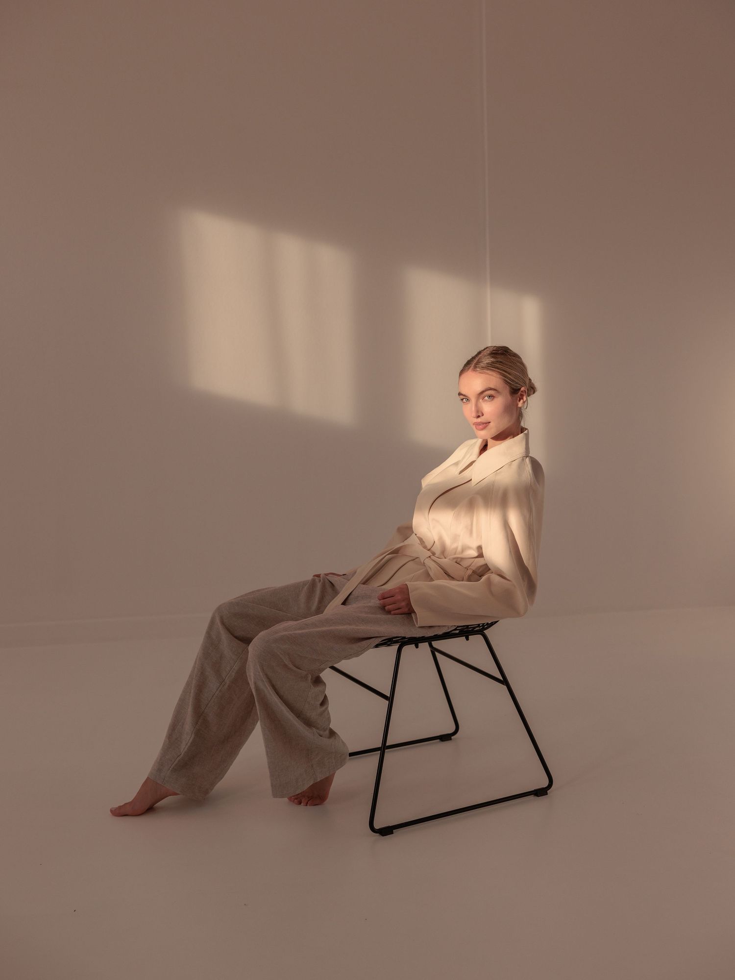 Woman in cream shirt and beige trousers sits on a black chair in a sunlit minimalist studio.