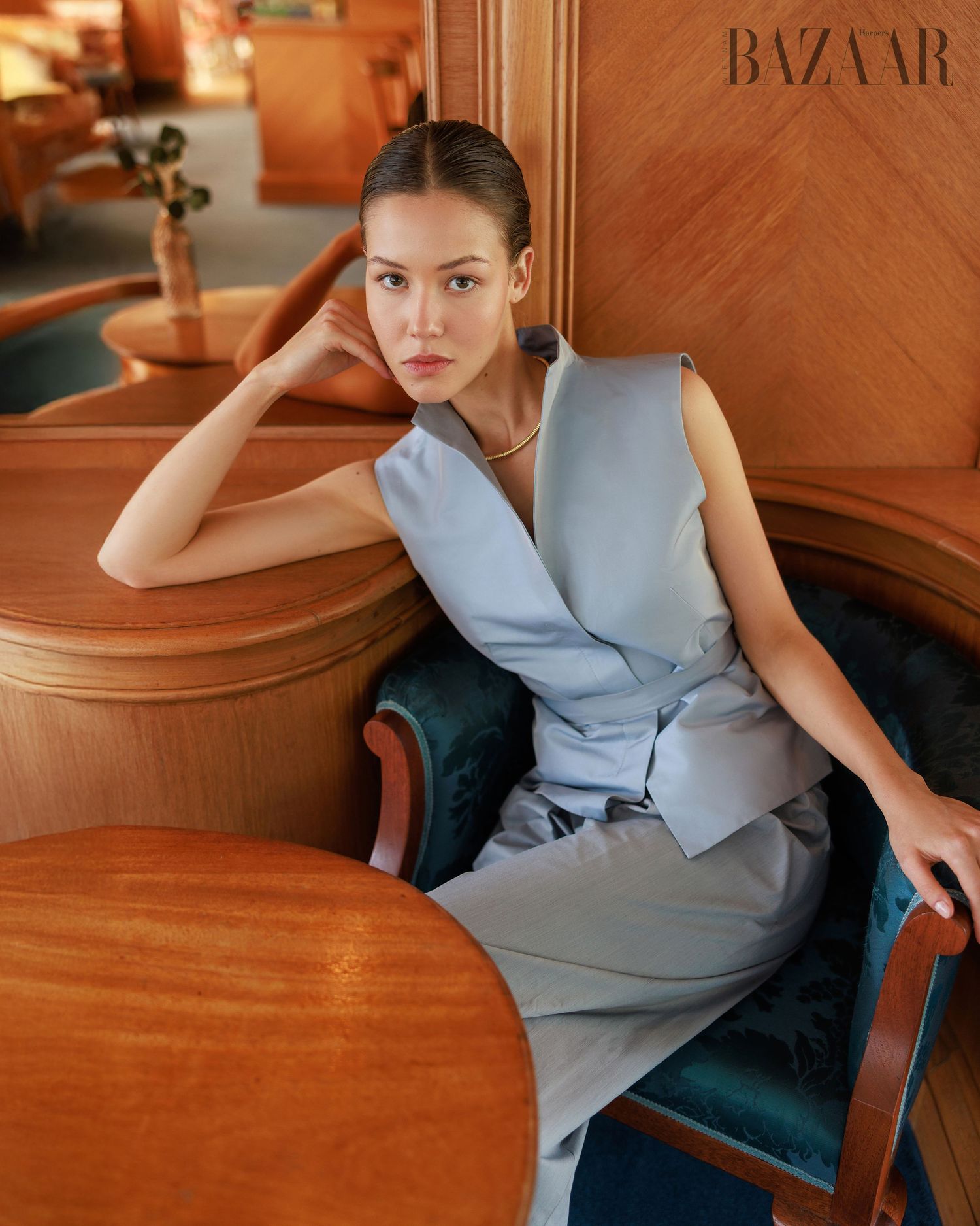 Model in light blue sleeveless belted vest and trousers, seated in warm wood-paneled interior for Harper's Bazaar.
