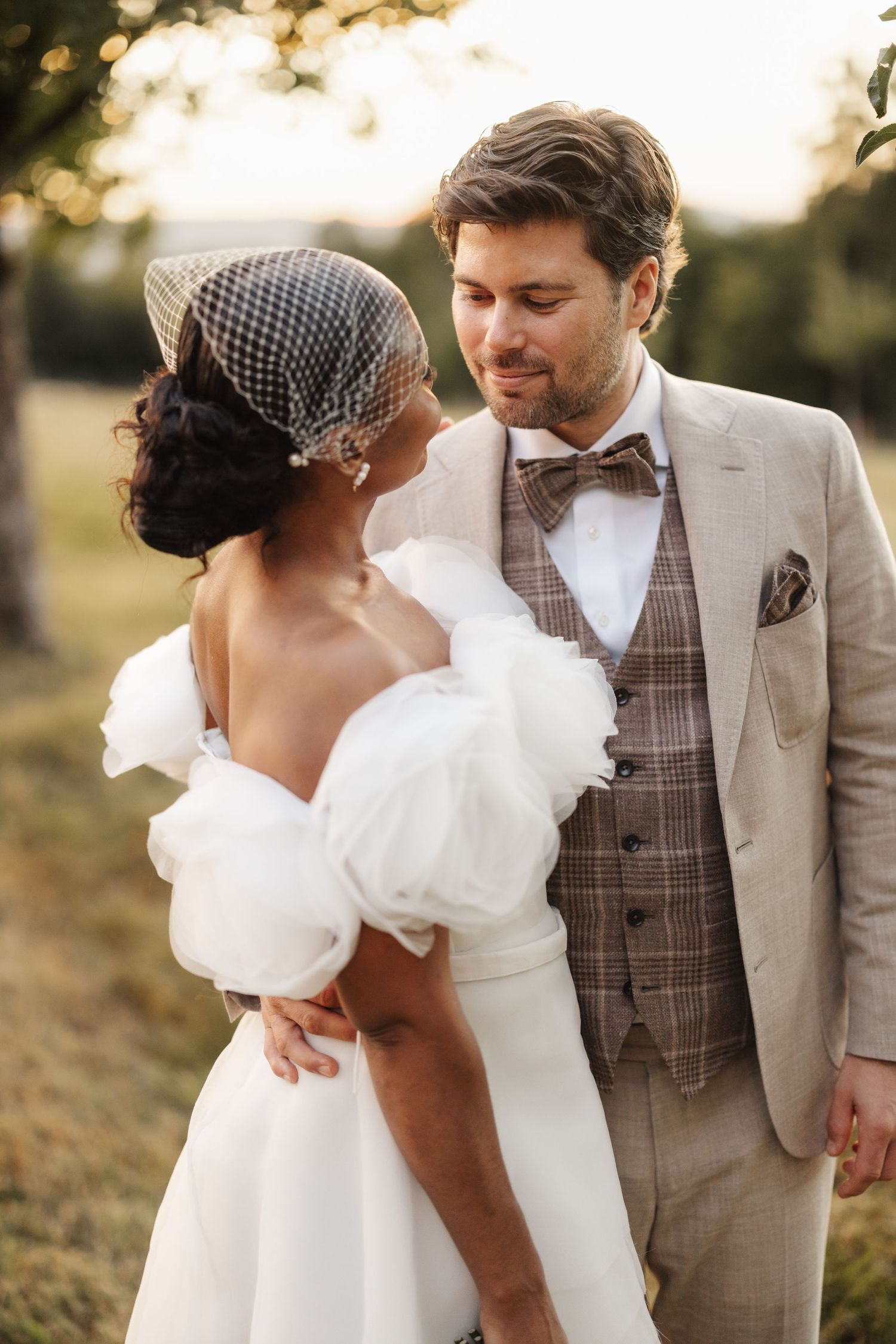 Groom in tweed vest and bow tie smiling at bride wearing off-shoulder white gown with birdcage veil outdoors.