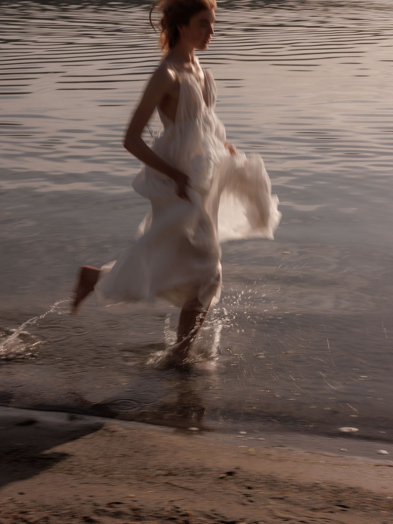 Woman in flowing white dress running through shallow water at golden hour, dress billowing in motion.