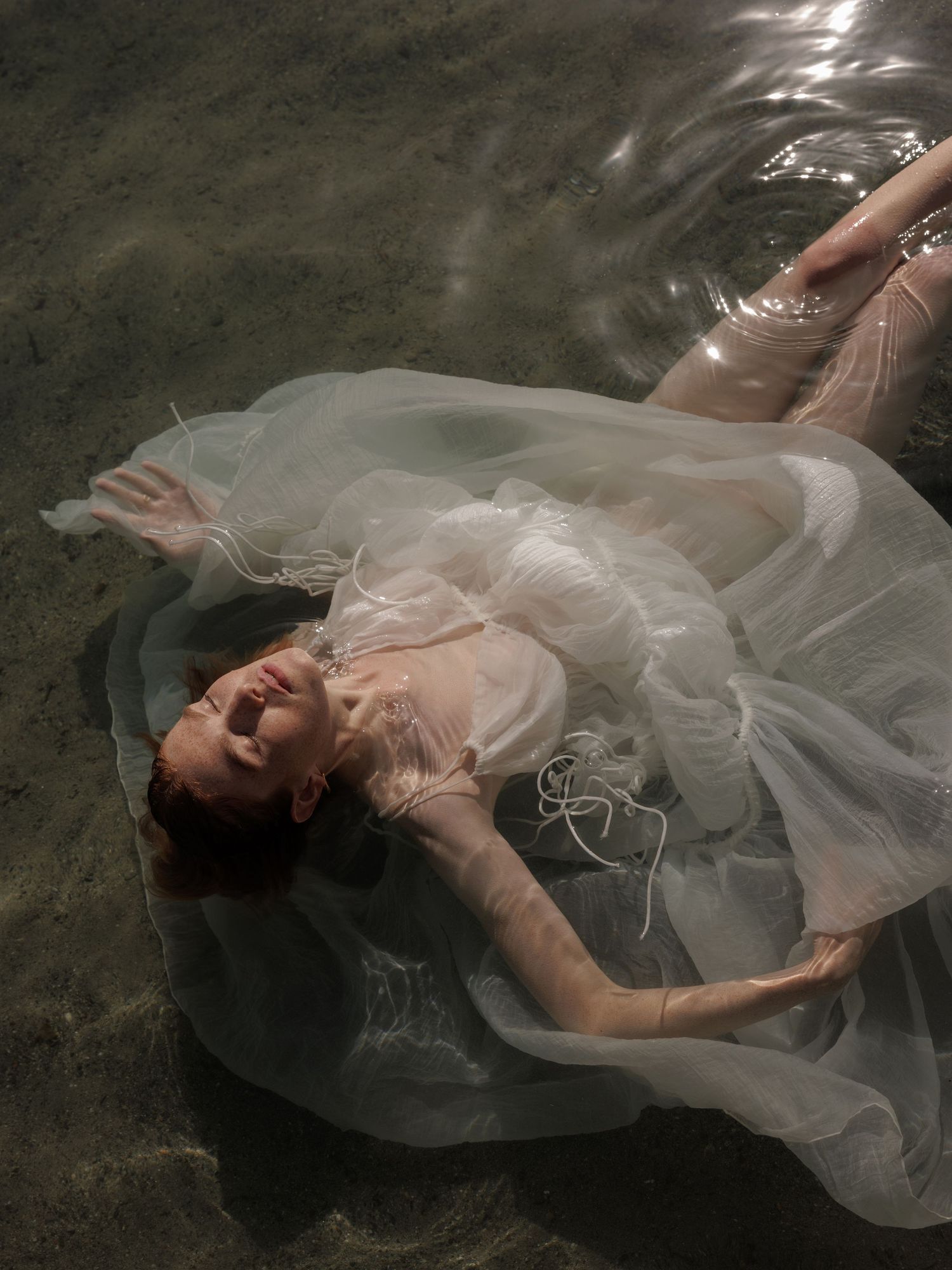 Woman in wet white tulle dress floating in shallow water, head tilted back with eyes closed.