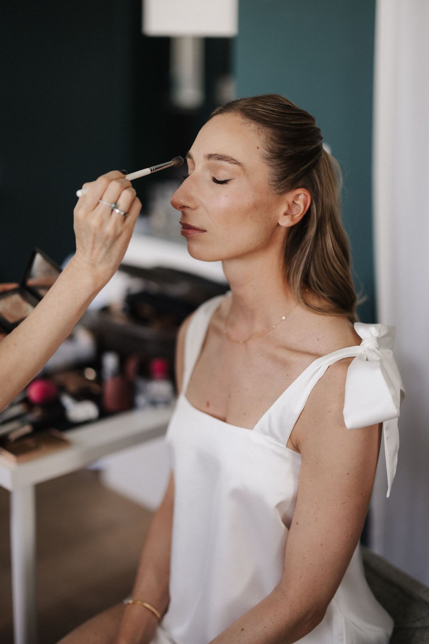 Makeup artist applying blush to a young woman in a white dress with bow straps.