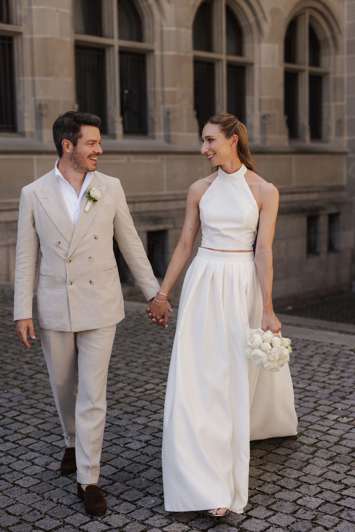 Happy bride and groom holding hands walking on cobblestone plaza, she carries white peonies, he wears beige double-breasted suit.