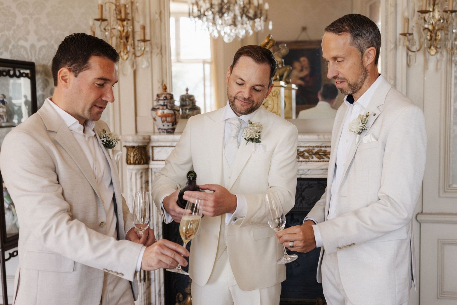 Groom and groomsmen in cream suits sharing champagne toast before wedding ceremony indoors.