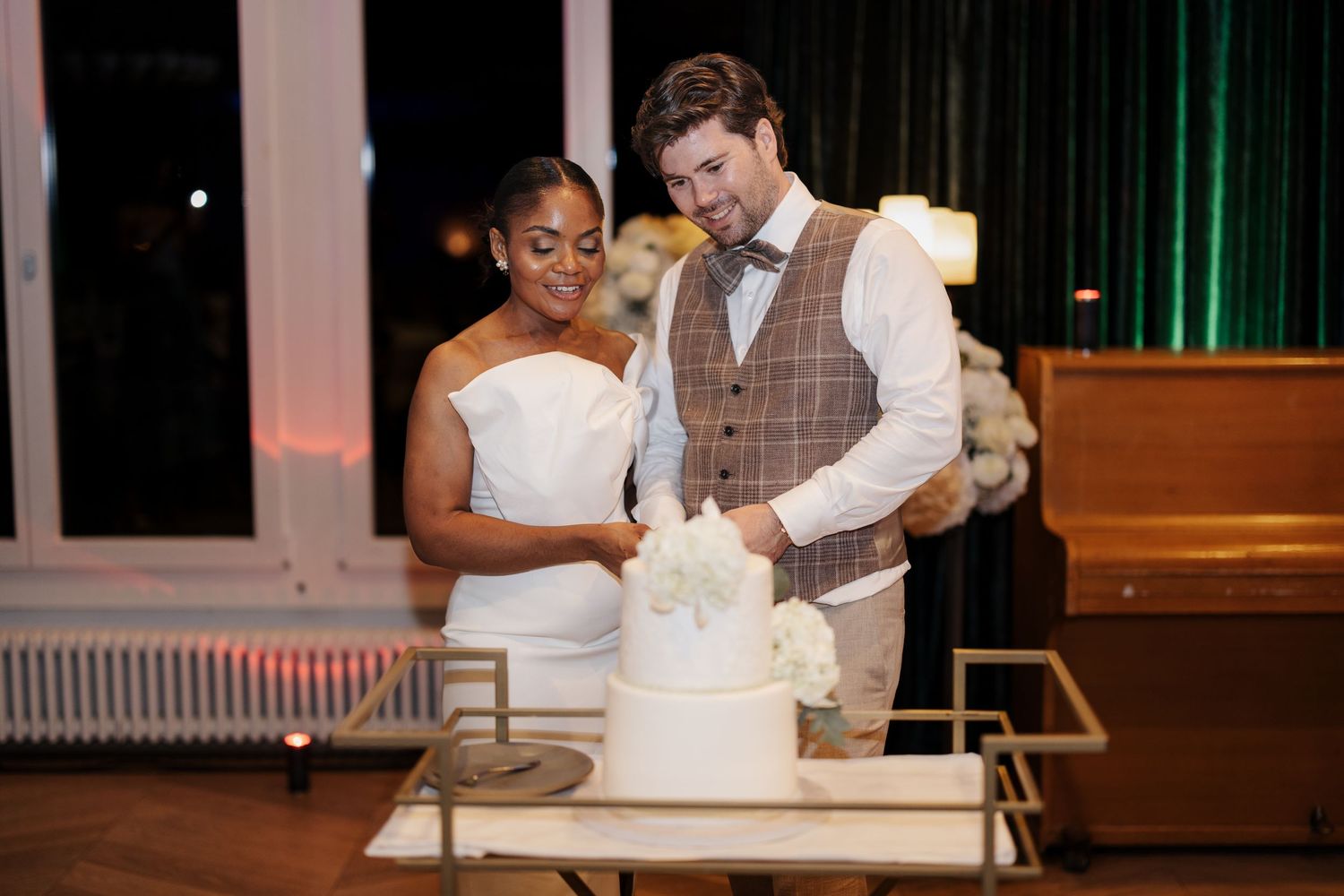 Interracial couple cutting their white tiered wedding cake at a romantic evening reception venue.