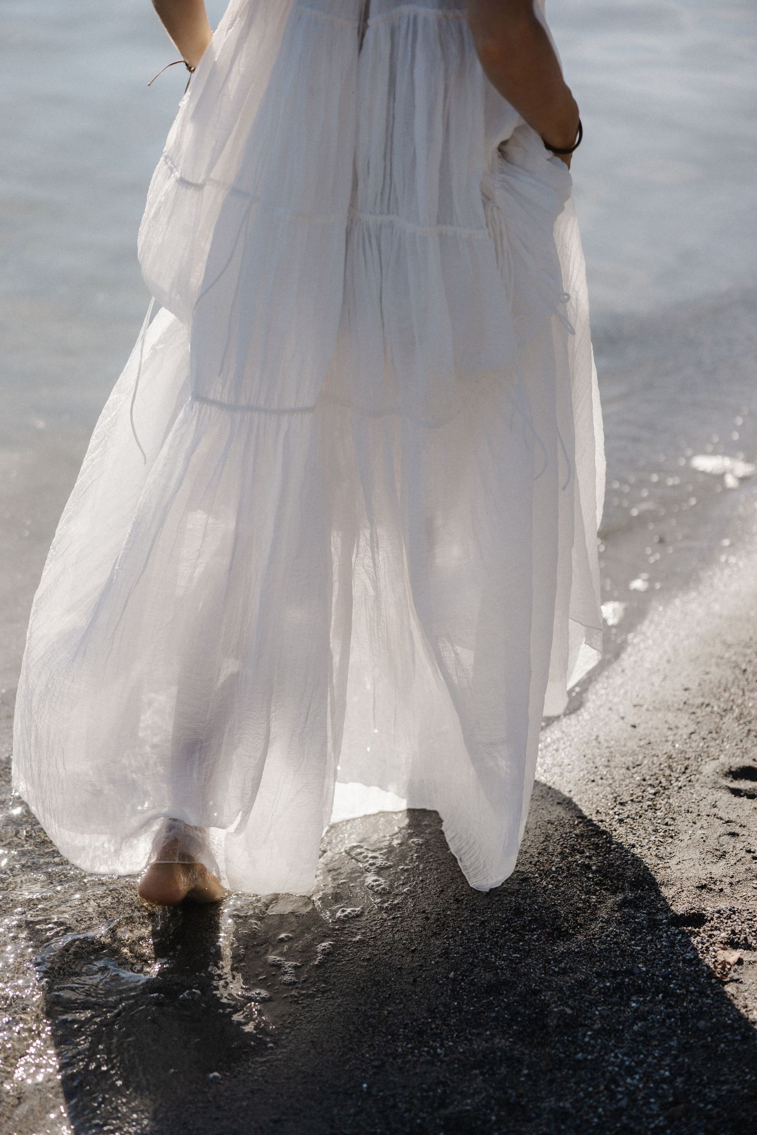 Woman in flowing white maxi dress walking barefoot along a wet sandy shoreline.