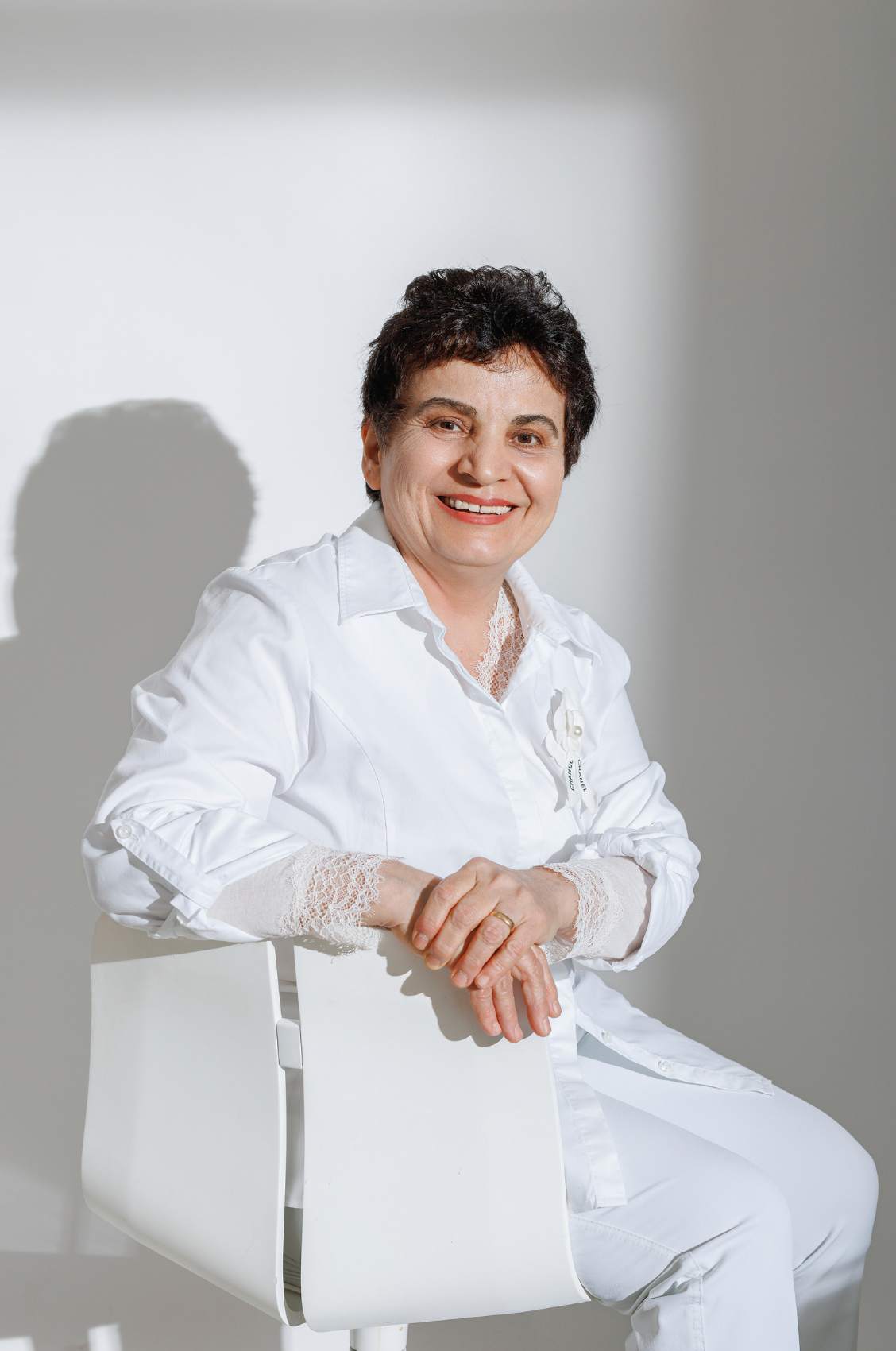 Mature woman in white outfit sitting on white chair, smiling warmly against light background.