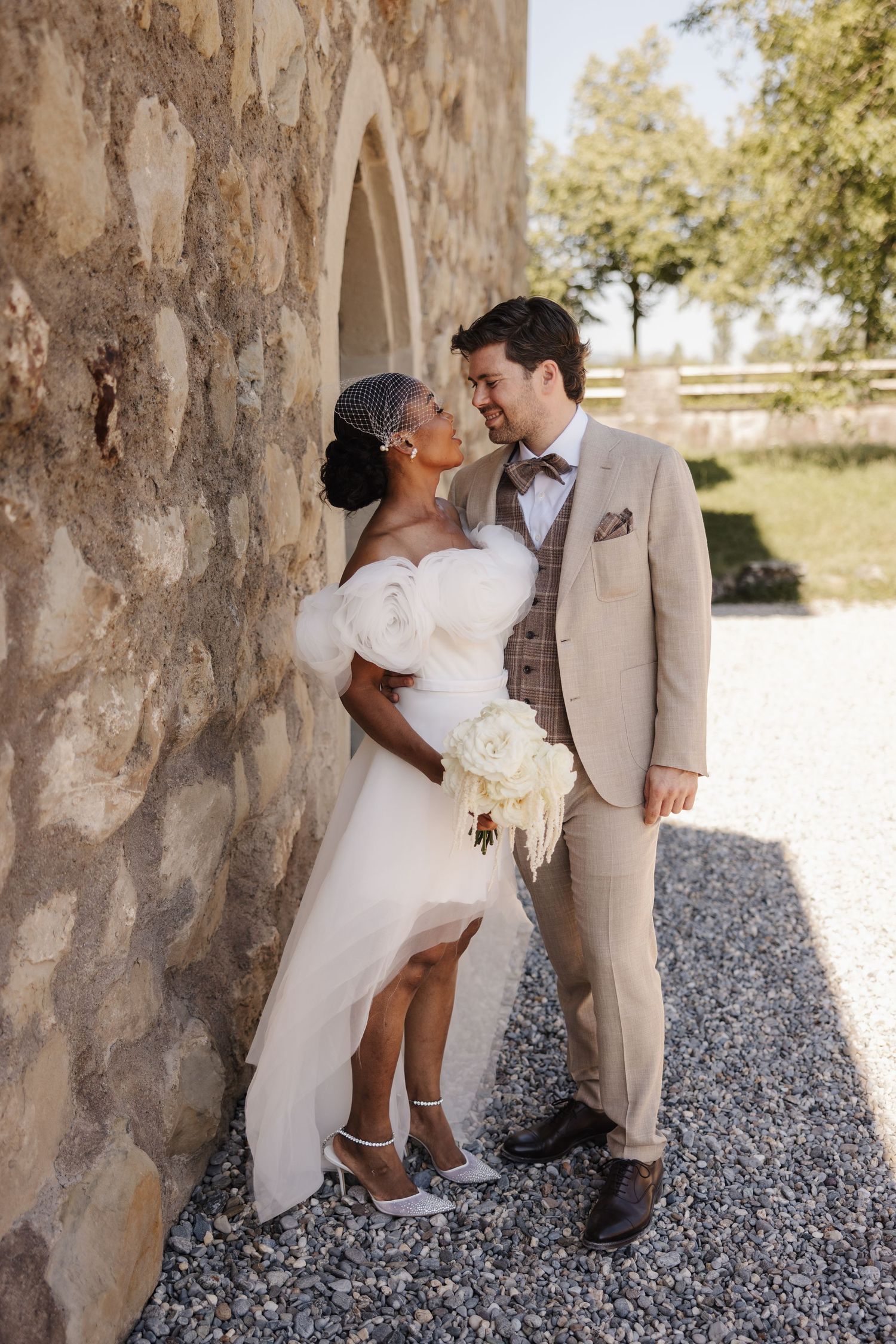 Romantic interracial newlywed couple leaning against a stone wall with a white bridal bouquet.