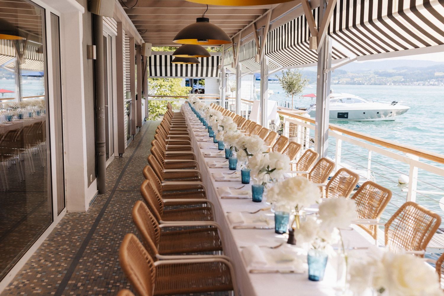 Waterfront restaurant terrace with long banquet table set for a wedding reception overlooking a lake with boats.